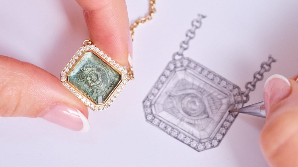 Hand holding a gold pendant with green and blue gemstone next to its outline drawing.