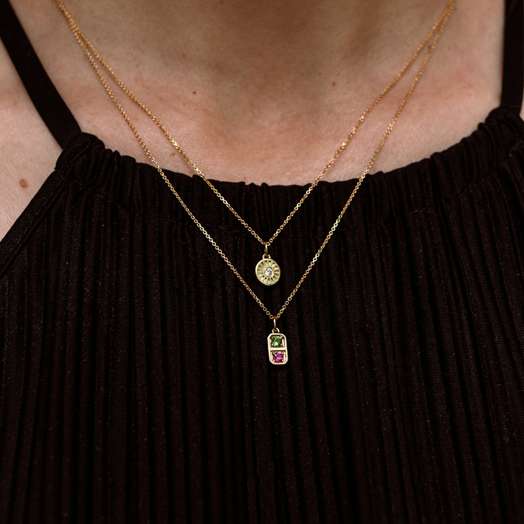 A close-up of a model wearing two layered gold necklaces. The shorter necklace features a round bezel-set crystal pendant, while the longer necklace hangs below it to show a small rectangular pendant with a green top and a pink bottom.