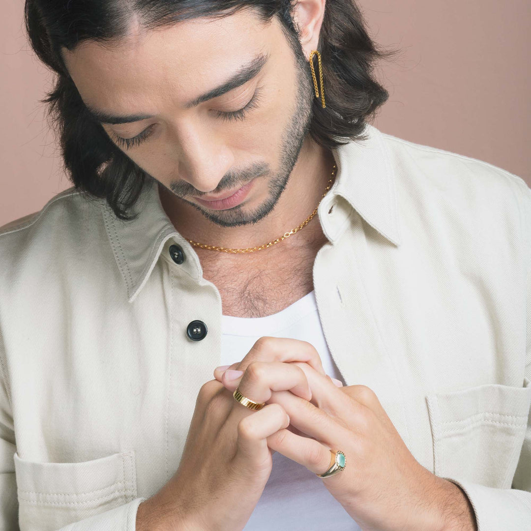 close up of man wearing gold chain earrings and necklace and looking at hands wearing gold diagonal groove male engagement ring and chrysoprase signet ring