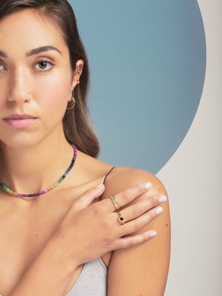 close up of woman wearing yellow gold rings and rainbow sapphire beaded necklace in front of a blue circle