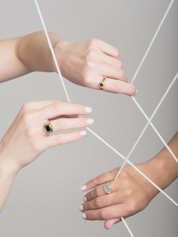 three hands holding onto white strings and wearing yellow gold hero rings set with gemstones