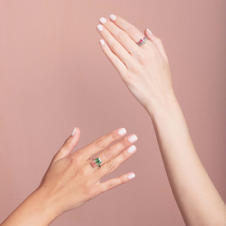 two womens hands enter from the left and right of this image, both are wearing yellow gold watermelon tourmaline rings. Set against a pink background